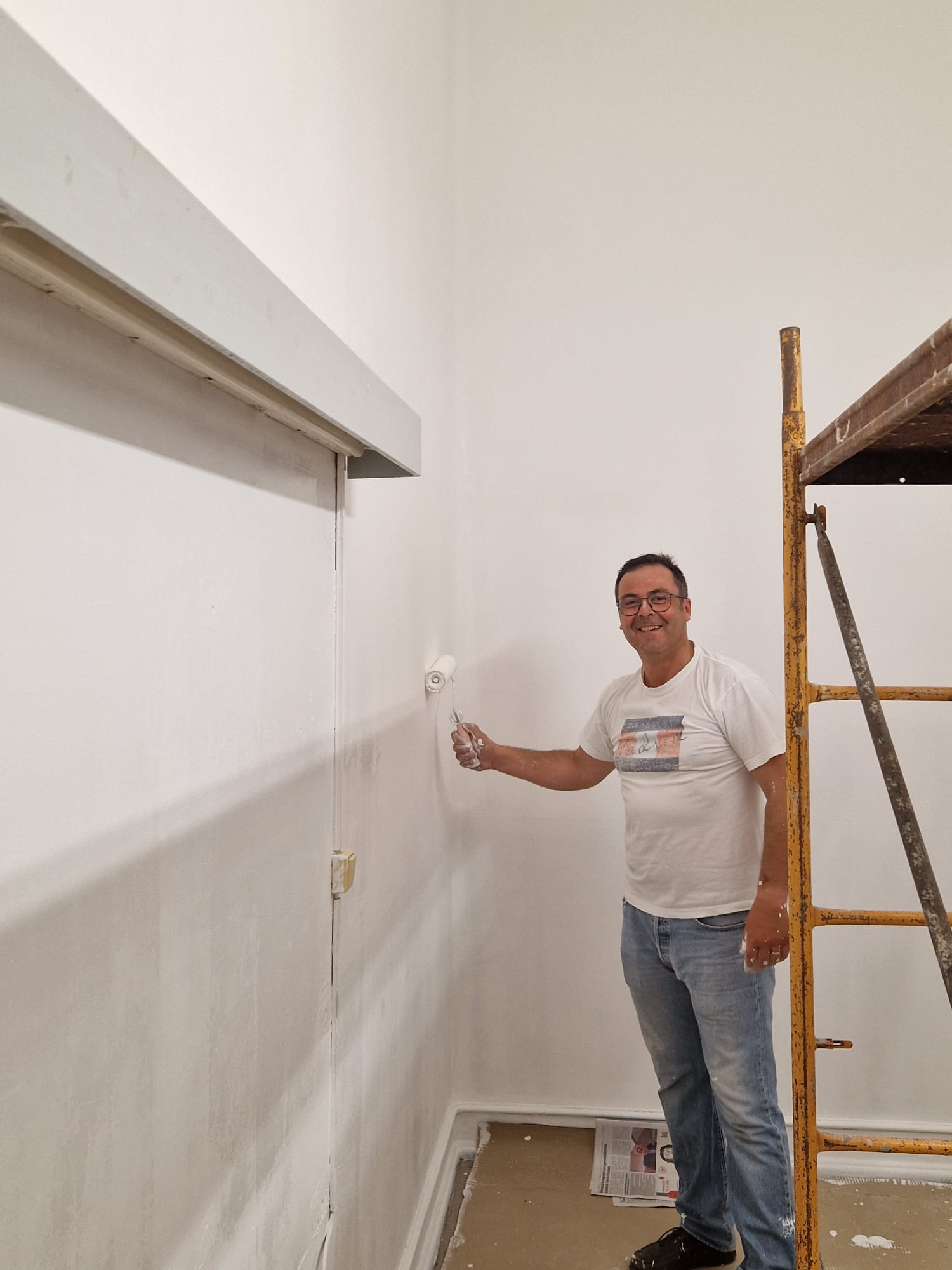 Um homem está a pintar uma parede branca com um rolo, dentro de uma sala. Ele está a sorrir para a câmara, usa óculos, t-shirt branca com uma estampagem retangular no peito, calças de ganga e sapatos escuros. Na mão direita ele segura o rolo com tinta branca. À direita da foto há um andaime metálico amarelo/enferrujado. No chão, há papelão e folhas de jornal protegendo a área, e dá para ver rodapés brancos ao longo da parede.